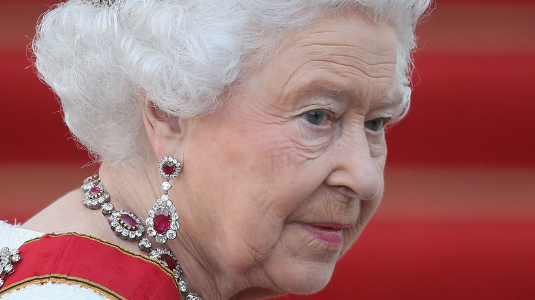 Queen Elizabeth wearing a crown, rubies, and diamonds standing on a red carpet 