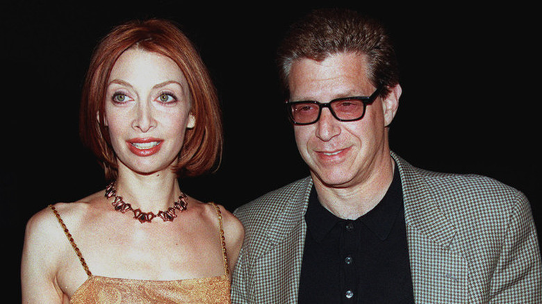 Actor Illeana Douglas looks off camera smiling wearing a necklace and red lipstick walking next to her ex-husband  Jonathan Axelrod who wears a checkered suit jacket