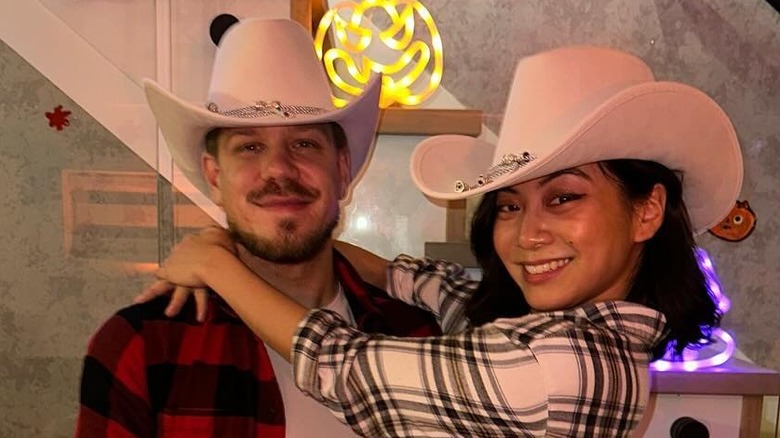 Actor Kallie Hu smiles wearing a cowboy hat with her arms wrapped around her smiling fiancé's neck as he holds her