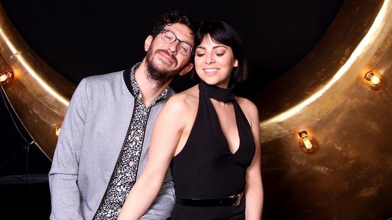 Actor Krysta Rodriguez wears a black tank top and smiles with her eyes closed next to her husband who also smiles with his eyes closed wearing a gray jacket