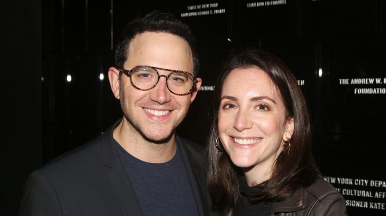 Santino Fontana wears glasses and stands next to his wife Jessica Hershberg smiling in front of a black sign