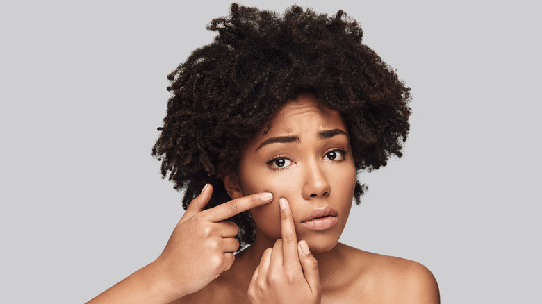 Woman with dark curly hair and bare shoulders with worried look on her face squeezes her cheek with both pointer fingers