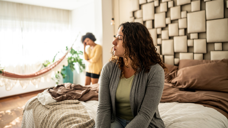 Woman sits on bed while other woman stands in background