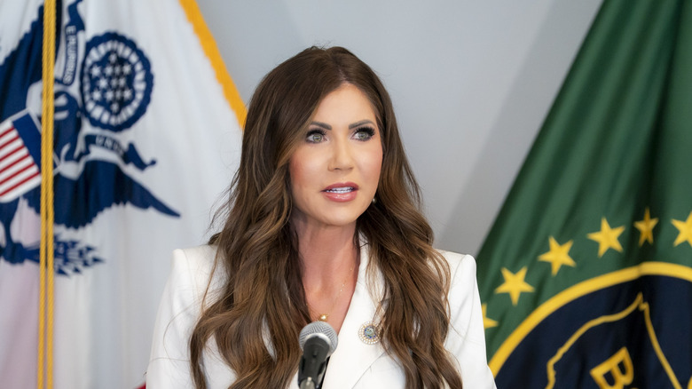 Kristi Noem speaking at a microphone in a white outfit
