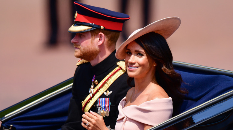 Meghan Markle smiling with Prince Harry while both wearing hats in an open-top car