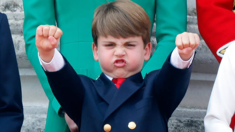 Prince Louis puffing his cheeks while fist pumping the air with both hands in a navy suit and red tie