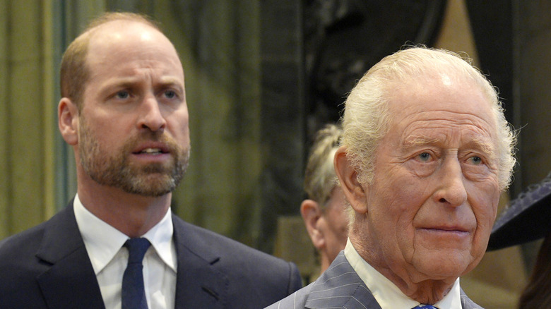 King Charles II standing in front of his eldest son, Prince William, while both wear dark grey suits