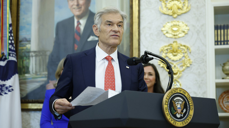 Dr. Oz giving a speech while holding papers in a navy suit with a red tie