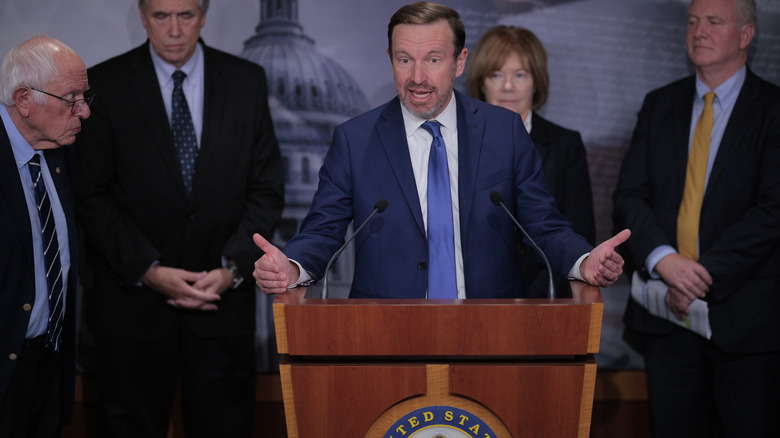 Chris Murphy delivering a speech while gesturing with his hands in a dark blue suit with a light blue tie