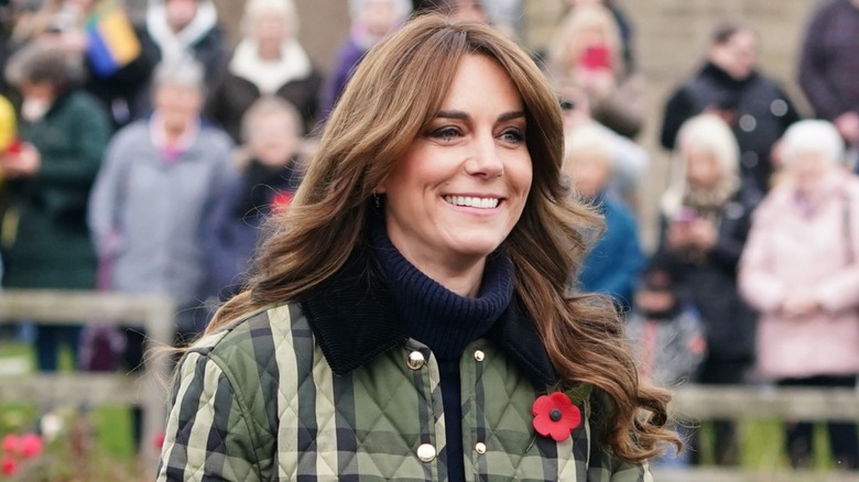 Kate Middleton wearing a green plaid jacket with a black turtleneck and poppy pin