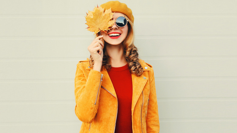 Woman holding yellow fall leaves covering one of her eyes and a yellow suede motocross jacket with a rust shirt