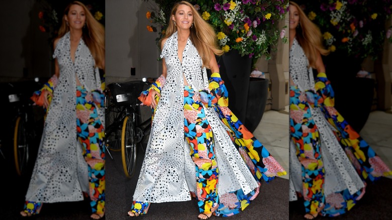 Blake Lively outside wearing a white blouse covered in black dots that goes to her ankles, big-flower patterned pants in blue, yellow and orange, and a matching long jacket