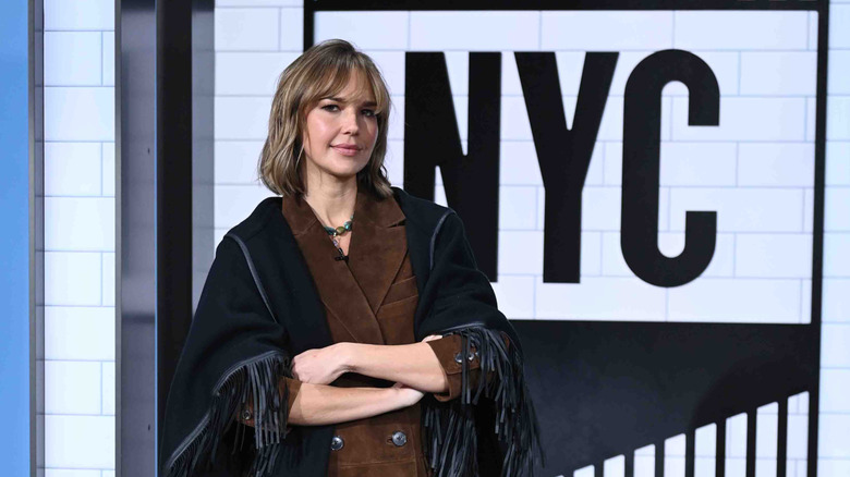 Arielle Kebbel posing in black and brown with her arms crossed on set with NYC written in the background