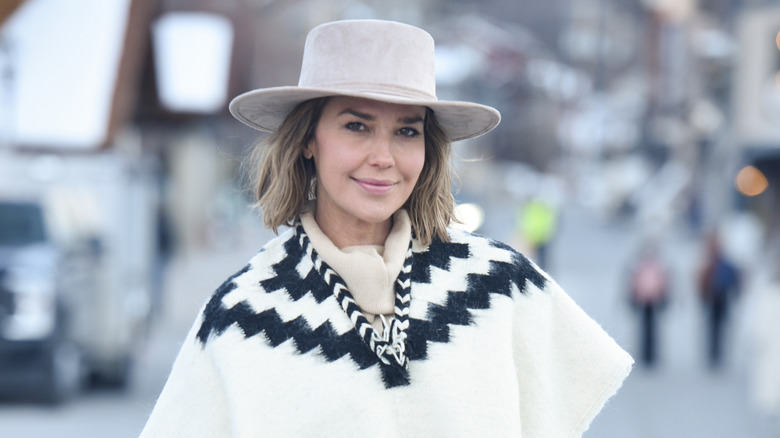 Arielle Kebbel wearing a beige hat and white and black sweater outside and smiling