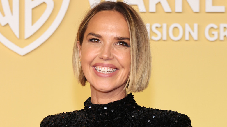 Arielle Kebbel smiling with a bob in front of a yellow background wearing a black sparkle turtleneck dress