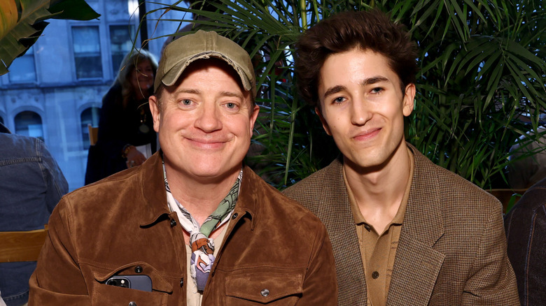 Brendan Fraser's son Holden smiles and sits next to him in front of a plant wearing a brown jacket