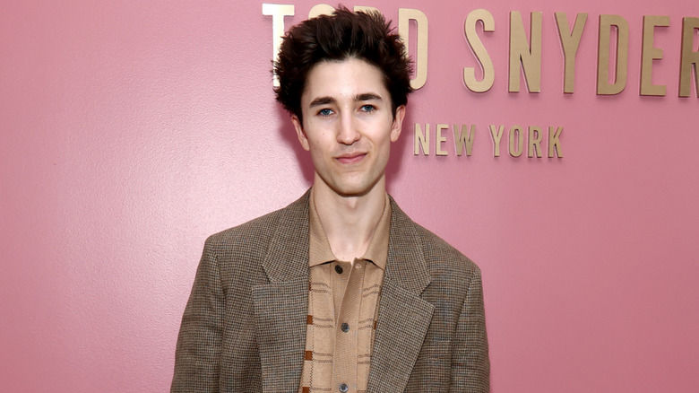Brendan Fraser's son Holden Fraser smiles in front of a pink wall at a fashion show wearing a brown jacket and shirt.
