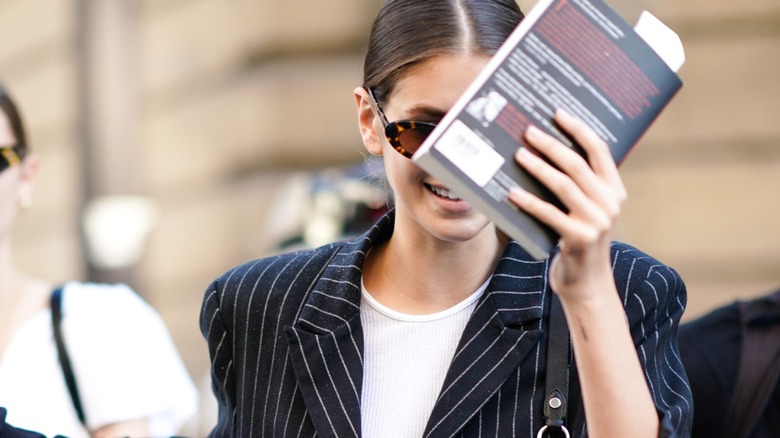 Kaia Gerber hiding her smiling face behind a book.