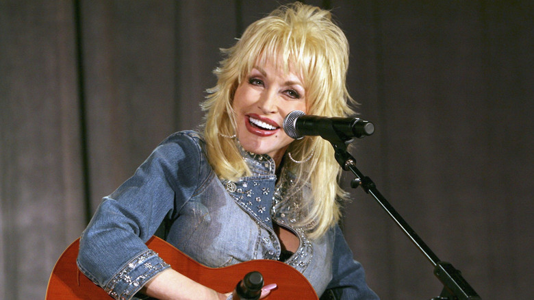 Dolly Parton smiles while playing a guitar and wearing a denim top