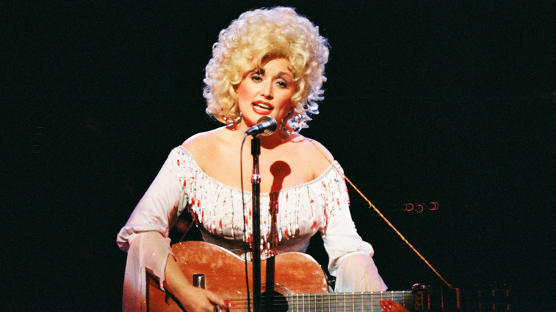 A young Dolly Parton plays an acoustic guitar and sings into a mic