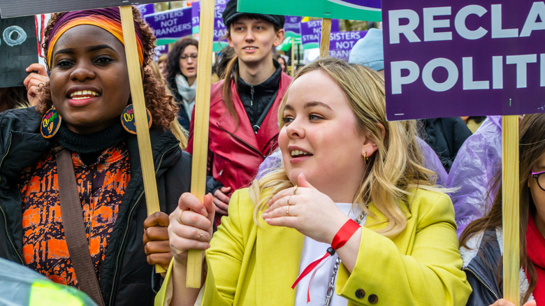 Nicola Coughlan in yellow blazer with red wristband holding a sign and marching in a protest