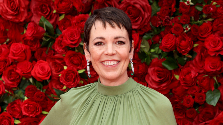 Olivia Colman smiles with short hair and bangs in front of roses wearing a green long sleeve dress
