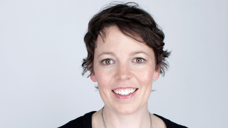Olivia Colman smiles against a white background in a black top with her short hair mussed in 2011