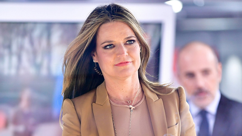 Savannah Guthrie walking on set in 2017 with earpiece with Matt Lauer blurred in the background