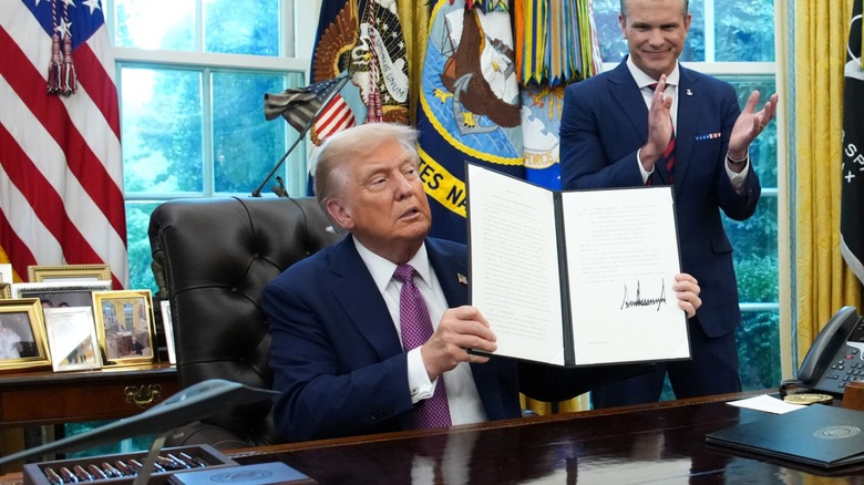Donald Trump holding up a signed executive order in the Oval Office, with Pete Hegseth clapping behind him