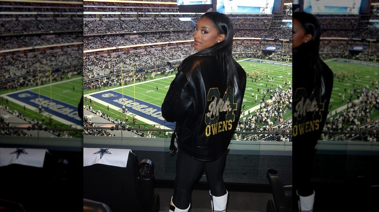 Simone Biles posing in front of a football field with a black jacket