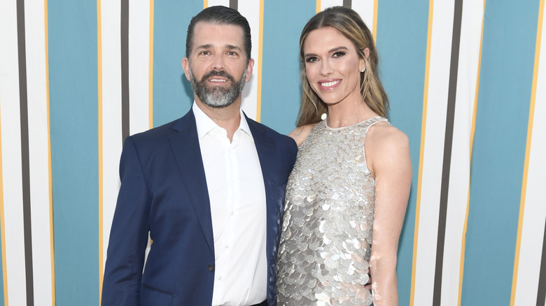 Donald Trump Jr. wearing a dark blue blazer and posing next to Bettina Anderson, who is wearing a silver dress covered with paillettes.