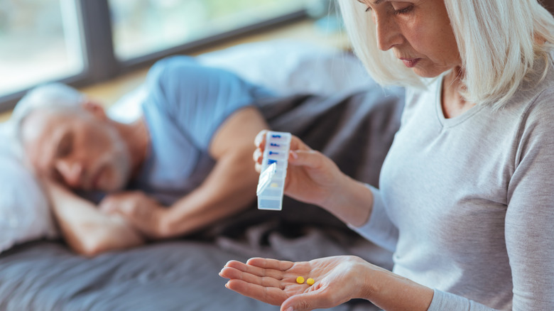 older woman taking medicine with husband sleeping in the background