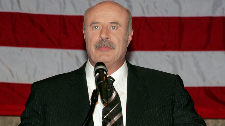 Phil McGraw wearing a black suit in front of a microphone and an American flag
