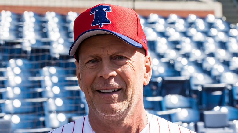 Bruce Willis smiling while wearing a baseball cap and uniform.