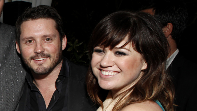 Kelly Clarkson in a black dress smiling next to Brandon Blackstock in a black suit.