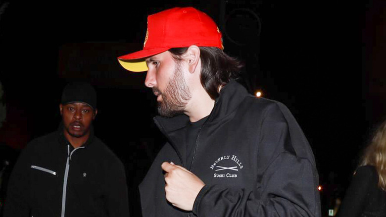 Scott Disick in profile, outside at night and wearing a red baseball cap and black windbreaker in 2022