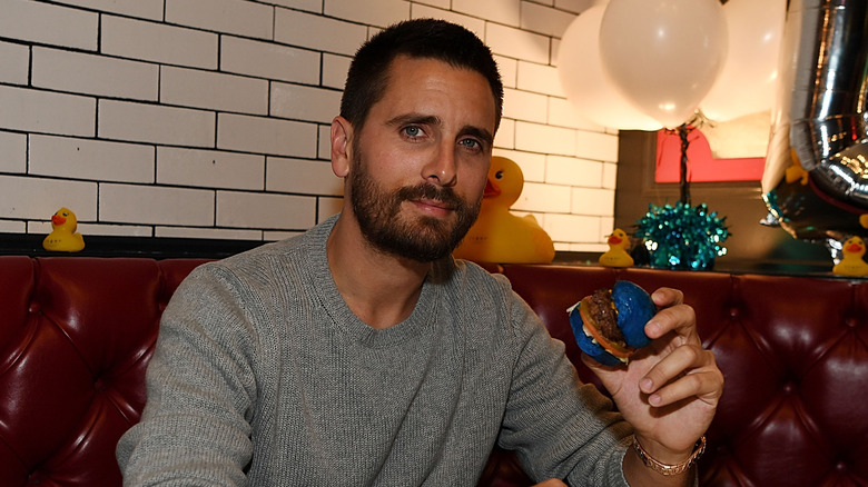 Scott Disick sitting in a red leather booth surrounded by rubber ducks and holding a tiny hamburger with a blue bun