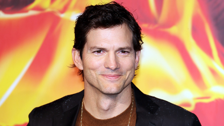 Ashton Kutcher smiling in a black blazer, brown sweater, and a gold chain