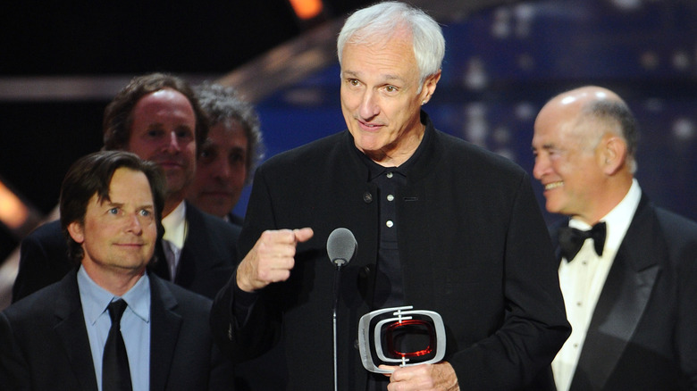 Michael Gross wears all black and stands next to Michael J. Fox in a suit onstage at the Fan Favorites Awards holding a trophy