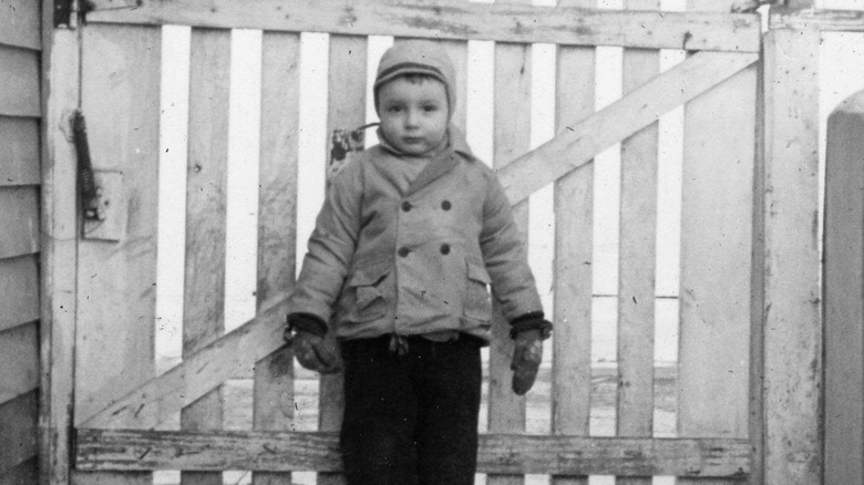 A young toddler Michael Gross stands in a black and white photo bundled up in winter in front of a fence