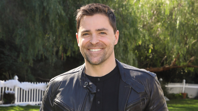 Kavan Smith in a black leather jacket smiling in a yard surrounded by trees and a white fence.