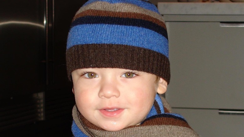 Joaquin Consuelos as a toddler smiling, while wearing a striped blue and brown winter hat and scarf