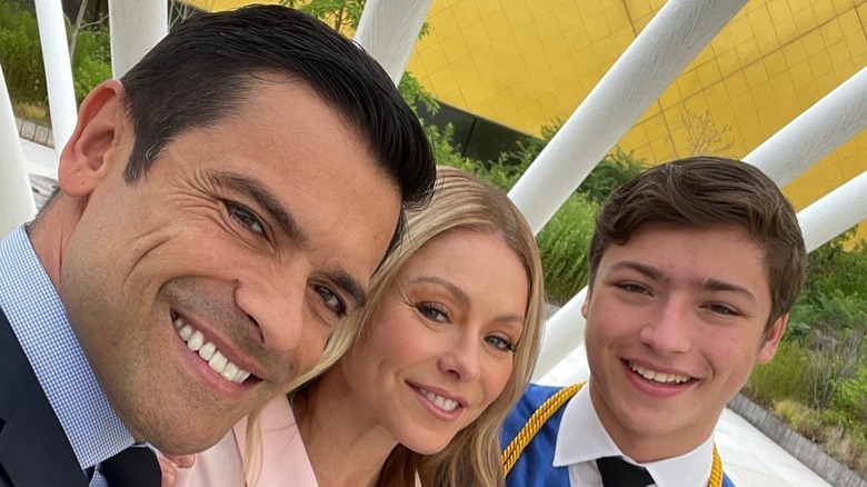 Joaquin Consuelos smiling in a selfie next to his parents on his high school graduation wearing a blue robe