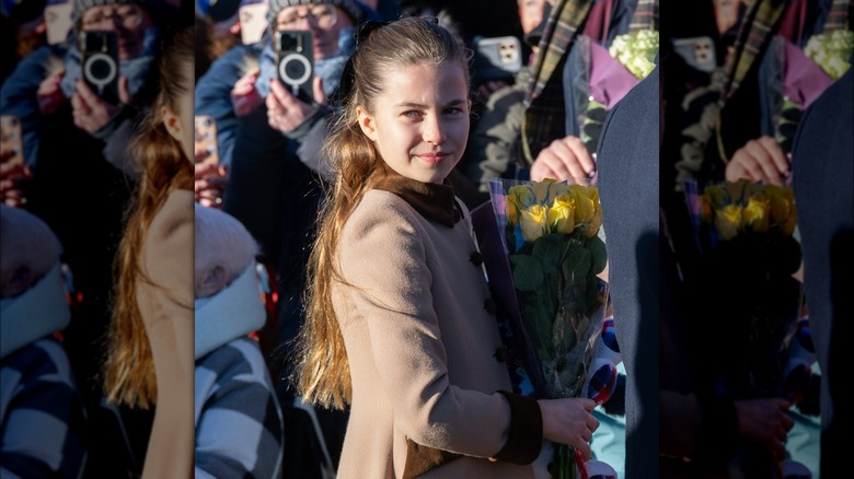 Princess Charlotte walking in a brown coat carrying a bouquet of yellow roses