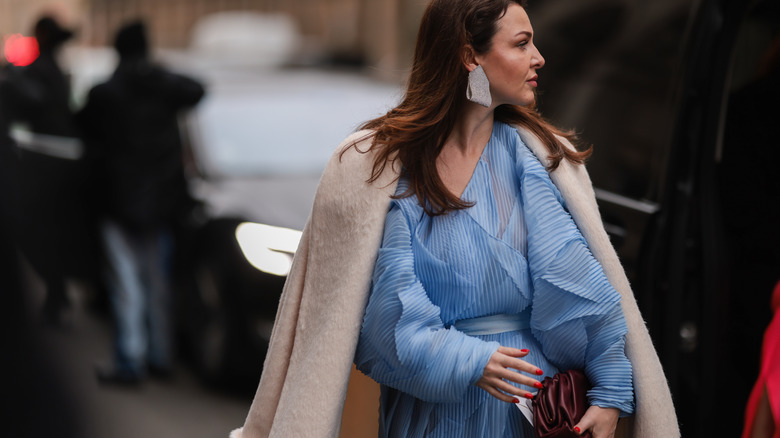 woman wearing winter 2022's trending colors through silver earrings, a brown jacket, blue dress, pink nails, and a purple handbag