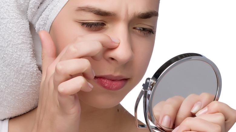 Woman wearing bath towel on her head and holding mirror points to her nose