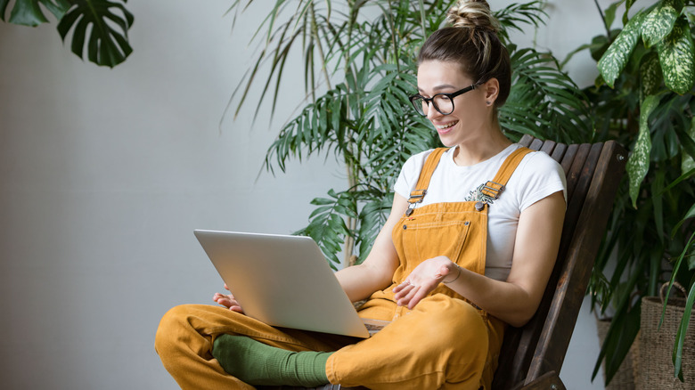 Woman wearing overalls on laptop surrounded by plants