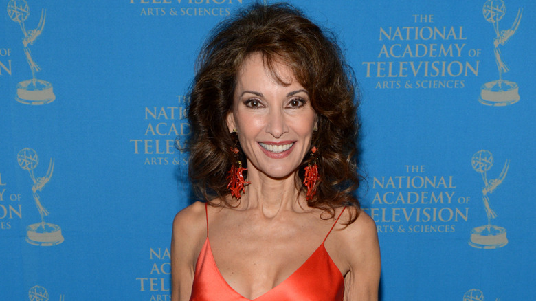 Susan Lucci smiling while posing in front of an Emmys backdrop in a strappy orange dress