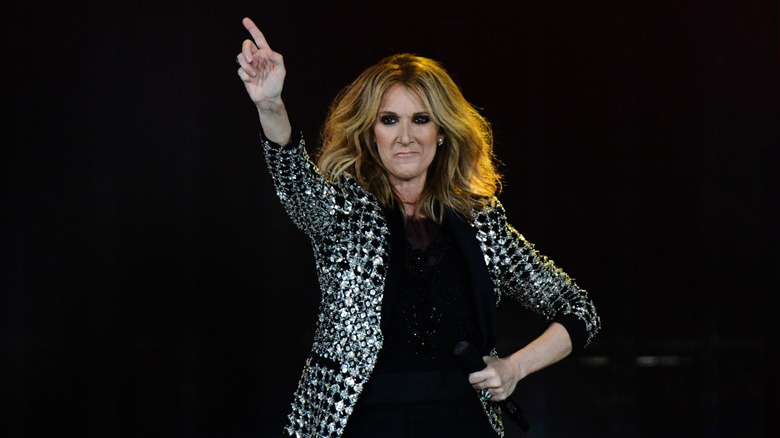 Céline Dion pointing toward the sky with her other hand on her hip