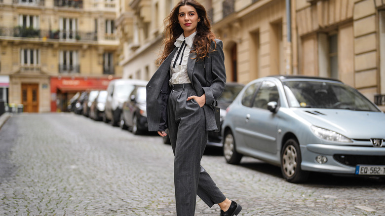 Giorgadze @katie.one wears gray tailored tapered pinstripe wool trousers from &OtherStories, a gray oversized asymmetric pinstripe blazer jacket from &OtherStories, classic masculine black leather loafers from Prada, a white ruffled emrboidered "broderie anglaise" cotton shirt with a black ribbon